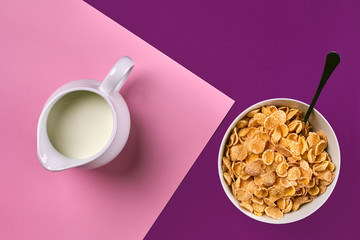 Obraz na płótnie Canvas Bowl with corn flakes, jug of milk and spoon on purple and pink background, top view