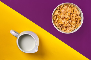 Obraz na płótnie Canvas Bowl with corn flakes, jug of milk on purple and yellow background, top view