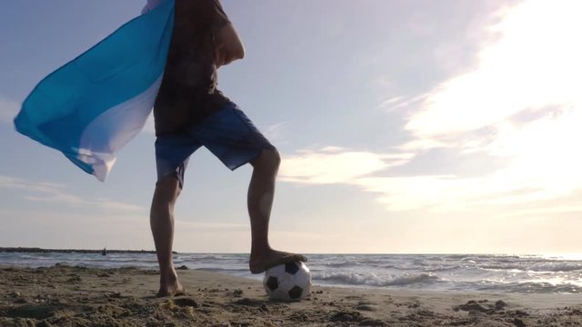 Young man wearing argentinian argentina national flag as super hero cape stands with foot on football on the sea shore sand looking at the ocean at the beach at sunset camera steadycam gimbal