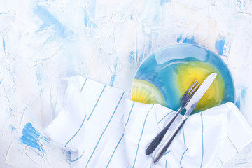 White table with colored blue. A white towel with blue stripes. A beautiful plate with a fork and knife. Free space for text, postcards or advertisements.