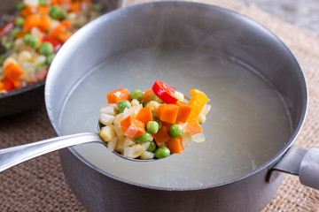 Mix vegetables on a spoon in a saucepan with boiling broth on a wooden table