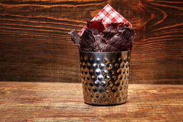 Dried peppered beef jerky cut in strips in a metal bucket on wooden background. Serving the dish in...