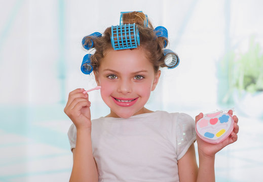 Close up of little girl using make up while wearing hair-rollers and bathrobe in a blurred background