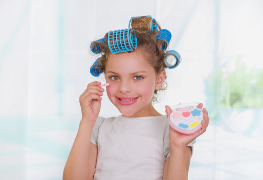 Close up of little girl using make up while wearing hair-rollers and bathrobe in a blurred background