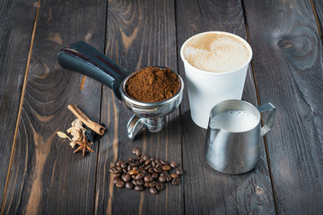 ingredients for coffee/ ingredients for coffee of Holder with ground coffee, spices and spices, coffee beans, cappuccino in a paper cup and a pitcher with milk on a wooden table.