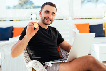 Young handsome man with laptop pointed bitcoin on camera sitting on deck at beack cafe on summer vocation. Man check bitcoin value on summer vocation.