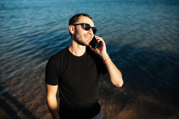 Handsome young man in sunglasses talking with mobile phone on the sea beach