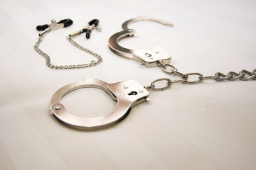 Fototapeta na wymiar handcuffs on the hands and feet on a chain with nipple clips against a white sheet