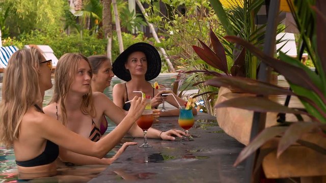 Beautiful caucasian women in swimsuits relaxing at outdoor swimming pool bar with colorful cocktails