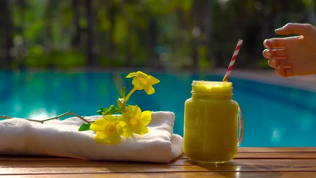 Hand of a woman puts a mango smoothie on a chaise-longue with a pool at a background. Fruit smoothie - healthy eating concept. Close up of detox smoothie with mango