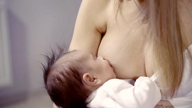 a little girl with brown hair is eating milk from her mother's breast, a lady is sitting with her bare chest
