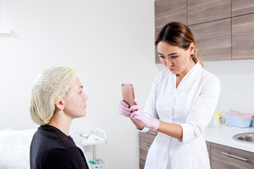 Woman azian doctor-cosmetologist takes pictures on the phone of the procedure performed by a cosmetology procedure on a young woman in a medical office