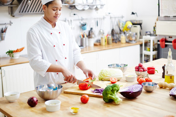 Young chef cutting fresh pepper with assortment of vegetables around on workplace
