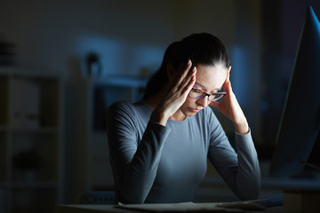 Young businesswoman with headache touching her head while sitting in front of computer monitor by her workplace at night