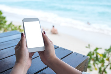 Mockup image of woman's hand holding white mobile phone with blank desktop screen on wooden table in front of the sea and blue sky background