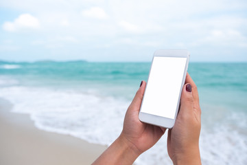 Mockup image of woman's hand holding white mobile phone with blank desktop screen in front of the sea and blue sky background