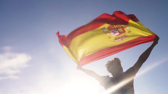 Young man holding spanish national flag to the sky with two hands at the beach at sunset spain