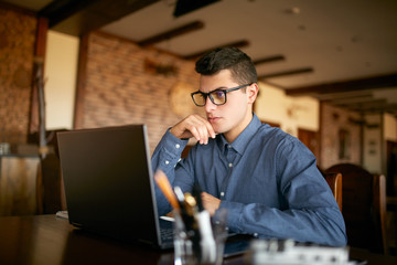 Thoughtful young caucasian businessman in glasses working on laptop computer. Pensive attractive...