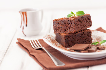 Chocolate brownie square pieces in stack on white plate decorated with mint leaves and cocoa powder on white vintage wooden background. American traditional delicious dessert. Close up photography