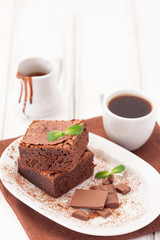 Chocolate brownie square pieces in stack on white plate decorated with mint leaves and cocoa powder on white vintage wooden background. American traditional delicious dessert. Close up photography