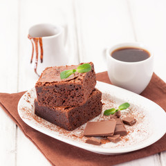 Chocolate brownie square pieces in stack on white plate decorated with mint leaves and cocoa powder on white vintage wooden background. American traditional delicious dessert. Close up photography