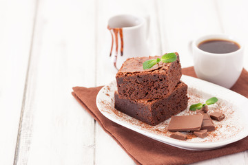 Chocolate brownie square pieces in stack on white plate decorated with mint leaves and cocoa powder on white vintage wooden background. American traditional delicious dessert. Close up photography