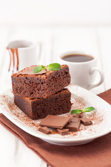 Chocolate brownie square pieces in stack on white plate decorated with mint leaves and cocoa powder on white vintage wooden background. American traditional delicious dessert. Close up photography