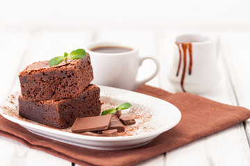 Chocolate brownie square pieces in stack on white plate decorated with mint leaves and cocoa powder on white vintage wooden background. American traditional delicious dessert. Close up photography
