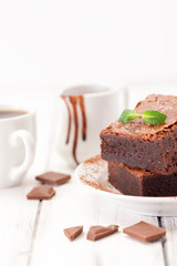 Chocolate brownie square pieces in stack on white plate decorated with mint leaves and cocoa powder on white vintage wooden background. American traditional delicious dessert. Close up photography