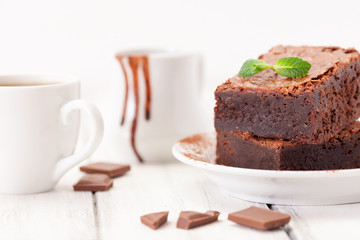 Chocolate brownie square pieces in stack on white plate decorated with mint leaves and cocoa powder on white vintage wooden background. American traditional delicious dessert. Close up photography