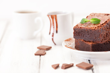 Chocolate brownie square pieces in stack on white plate decorated with mint leaves and cocoa powder on white vintage wooden background. American traditional delicious dessert. Close up photography