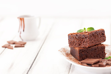 Chocolate brownie square pieces in stack on white plate decorated with mint leaves and cocoa powder on white vintage wooden background. American traditional delicious dessert. Close up photography