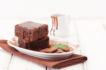 Chocolate brownie square pieces in stack on white plate decorated with mint leaves and cocoa powder on white vintage wooden background. American traditional delicious dessert. Close up photography