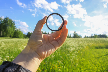 Hand holds a circular polarizer filter on summer landscape background.
