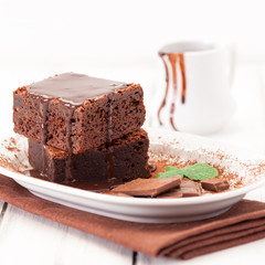 Chocolate brownie square pieces in stack on white plate decorated with mint leaves and cocoa powder on white vintage wooden background. American traditional delicious dessert. Close up photography