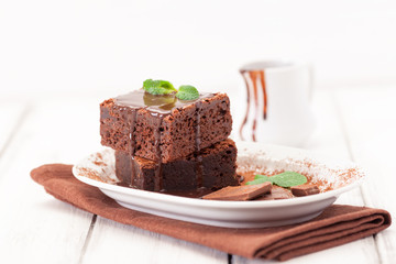 Chocolate brownie square pieces in stack on white plate decorated with mint leaves and cocoa powder on white vintage wooden background. American traditional delicious dessert. Close up photography