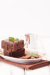 Chocolate brownie square pieces in stack on white plate decorated with mint leaves and cocoa powder on white vintage wooden background. American traditional delicious dessert. Close up photography
