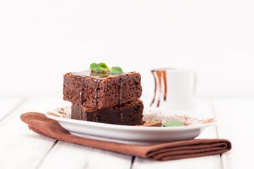 Chocolate brownie square pieces in stack on white plate decorated with mint leaves and cocoa powder on white vintage wooden background. American traditional delicious dessert. Close up photography