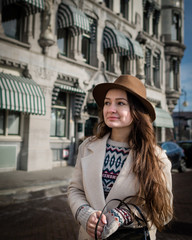 Portrait of a young female tourist with elegant clothes walking in the city. Trendy woman traveler with smile looking on buildings and touring old town. Beautiful girl exploring Rotterdam, Netherlands