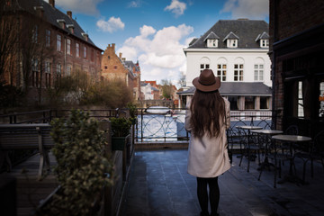 Fototapeta premium Young unrecognizable woman staying back in romantic place in the old town in Brugge, Belgium. Female tourist is inspired by atmosphere of vintage buildings on river promenade