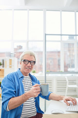 Grey-haired businessman with cup of coffee sitting by his workplace in office and having break