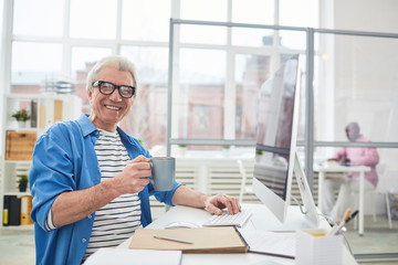 Smiling mature man in casualwear holdin gcup of coffee while sitting by desk in office