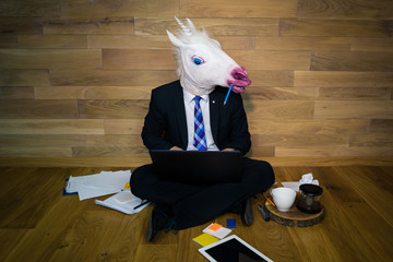 Young man in funny horse mask sits on the floor against a wall and works with laptop.  Serious unicorn in a suit and tie enthusiastically works at home with cup of coffee on wooden stand © Sergey_T