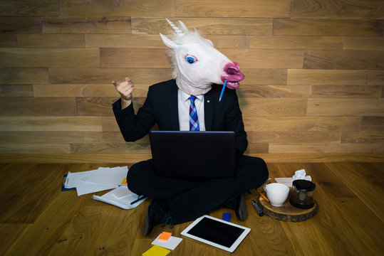 Motivator Unicorn Points To You! Young Man Sits On The Floor With A Coffee Near A Wall And Works In Laptop. Serious Guy In Funny Rubber Mask In A Suit And Tie Shows Thumbs Up At Home Office