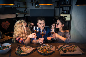 Three friends talks stories to each other in modern home kitchen at the bar counter. Group of attractive people has dinner together and holds glasses of wine. Young happy people enjoying time together