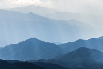 Scenic hazy blue mountains range covered by soft fog touching sunlight from cloudy sky.