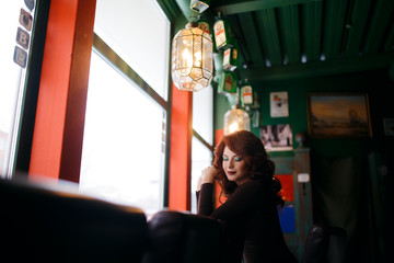 Thoughtful girl sits in cafe with red hair, wavy hairdo, red lips, green bureaucratic shadows in...