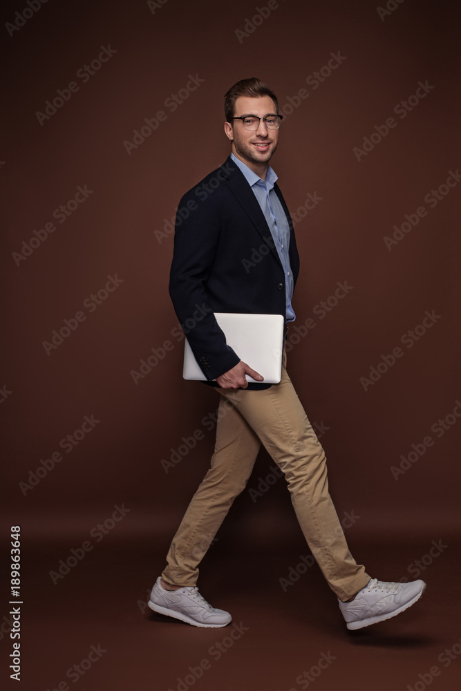 Poster full length portrait of happy businessman walking while holding notebook computer in arm. gadget and