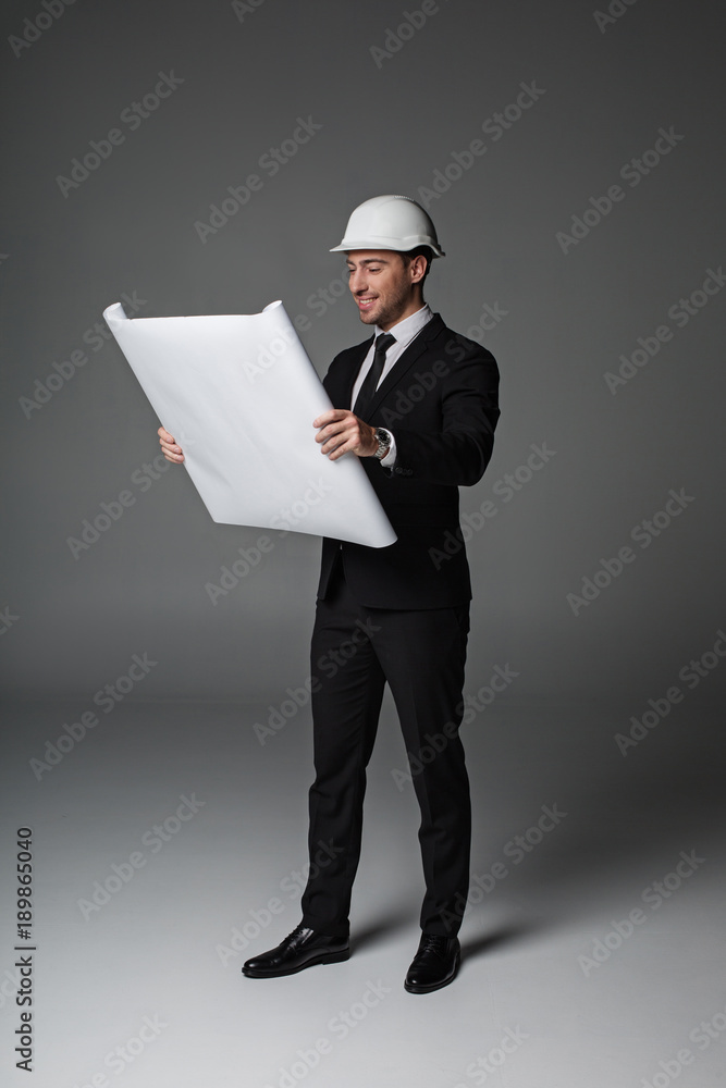 Poster full length portrait of cheerful unshaven architect looking at paper while keeping it in arms. const