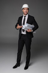 Full length portrait of beaming bearded master-builder pointing at plans by arm. Labor concept